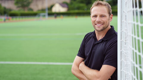 Mann lehnt lächelnd an einem Fußballtor auf dem Sportplatz