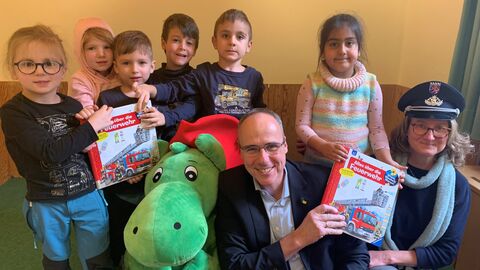 Innenminister Beuth kniet zwischen Kindern und einem grünen Plüschdinosaurier und lächelt in der Kamera, in der Hand hält er ein Buch.