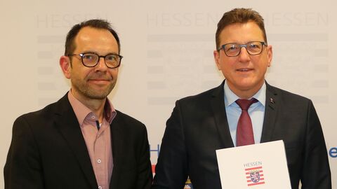 Dr. Reiner Becker steht neben Staatssekretär Sauer, dieser hält den Förderbescheid in der Hand. Beide lächeln in die Kamera.