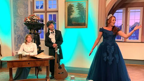 Eine Szene aus dem Theaterstück des IDRH: Eine Frau in blauem Ballkleid und mit Krone steht vorne, dahinter zwei Personen an einem Tisch sitzend.