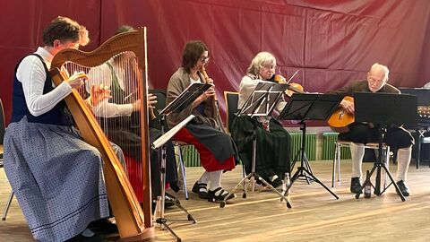 Vorweihnachtliche Egerländer Stubenmusik  Fünf Männer und Frauen sitzen auf einer Bühne und musizieren. Darunter ist eine Harfe, Blasinstrumente, eine Geige und eine Gitarre.