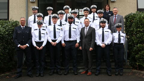 Die 25 neuen Wachpolizisten und Wachpolizistinnen stehen in Uniform zum Gruppenfoto auf einer Treppe vor einem Gebäude aufgestellt. In der ersten Reihe in der Mitte steht bei Ihnen Staatssekretär Rößler.