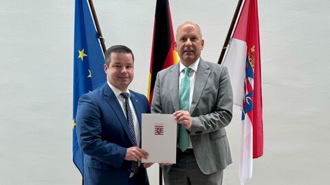 Andreas Hofmeister mit Innenminister Poseck Andreas Hofmeister (links) mit Innenminister Poseck (rechts). Die beiden stehen nebeneinander und halten gemeinsam ein Dokument in den Händen. Im Hintergrund sind die Flaggen des Landes Hessen, Deutschlands und der EU zu sehen.