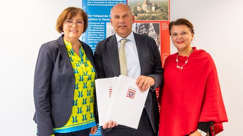 Innenminister Poseck (Mitte) steht neben zwei Frauen vor einem Aufsteller und hält die Förderbescheide in Händen.