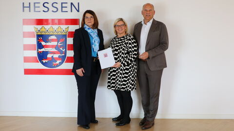 Innenminister Roman Poseck zusammen mit den Bürgermeisterinnen Henrike Strauch und Cäcilia Reichert-Dietzel vor Wand mit Hessen-Wappen