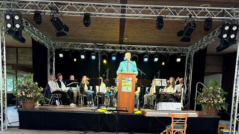 Staatssekretär Martin Rößler steht auf einer Bühne am Rednerpult und hält ein Grußwort, im Hintergrund sitzen Musiker.