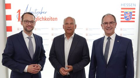 Drei Männer im Anzügen blicken in Kamera. Im Hintergrund sieht man das Hessen-Logo