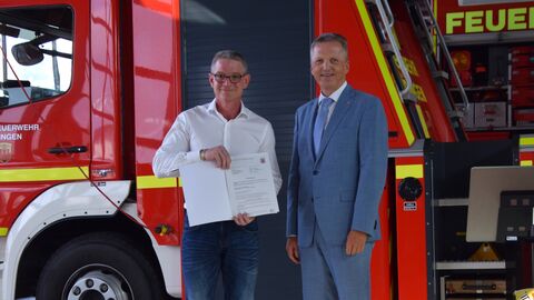 Staatssekretär Martin Rößler und Bürgermeister Benjamin Harris stehen vor einem Feuerwehrfahrzeug. Benjamin Harris hält den Zuwendungsbescheid in Händen.