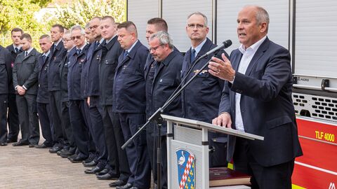 Innenminister Poseck steht an einem Rednerpult und spricht. Im Hintergrund ist ein Feuerwehrfahrzeug erkennbar. Neben ihm stehen Mitglieder der Feuerwehr.