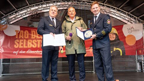 Innenminister Poseck steht mit zwei weiteren Personen auf einer Bühne und überreicht die Ehrenplakette.