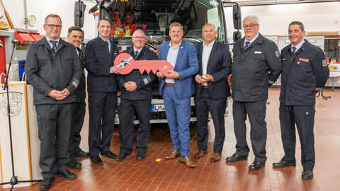 Innenminister Poseck steht mit Personen der Freiwilligen Feuerwehr Weilmünster in einer Halle. Im Hintergrund steht ein Feuerwerhfahrzeug. Zwei Personen halten einen große roten Schlüssel in Händen.