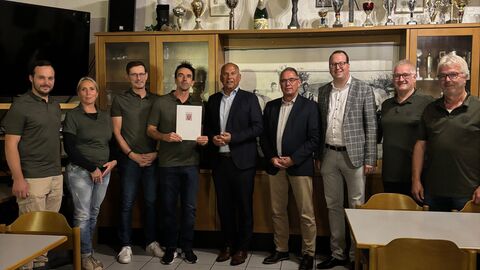 9 Menschen stehen nebeneinander in einem Vereinshaus.