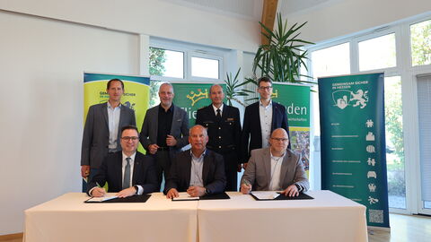 3 Männer sitzen an einem Tisch und unterzeichnen einen Vertrag. Hinter jedem steht jeweils eine weitere Person.