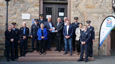 Innenminister Roman Poseck u.a. mit dem Bürgermeister von Alsfeld, Stephan Paule, bei der Überreichung des KOMPASS-Sicherheitssiegels. Alle stehen gemeinsam auf einer Treppe vor einem Gebäude.