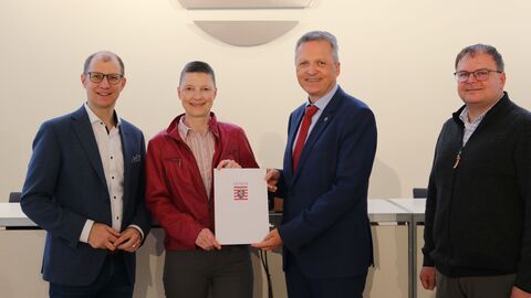 Staatssekretär Martin Rößler überreicht der Kreisbeigeordneten Marion Götz den Zuwendungsbescheid. Vier Personen halten ein Dokument in die Kamera und lächeln.