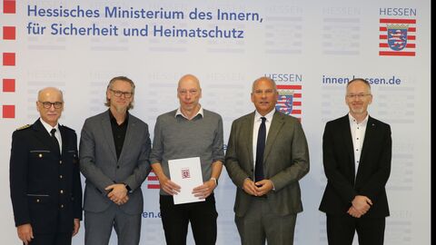 Innenminister Roman Poseck, Thomas Mücke vom Violence Prevention Network sowie 3 weitere Herren stehen vor einer Wand mit dem Logo des HdmI. Thomas Mücke hält den Zuwendungsbescheid in den Händen.