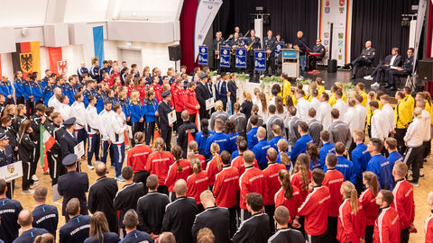 Viele junge Menschen stehen in Gruppen mit verschiedfarbigen Trikotjacken vor einer Bühne. Auf der Bühne steht Innenminister Roman Poseck und das Polizeiorchester.