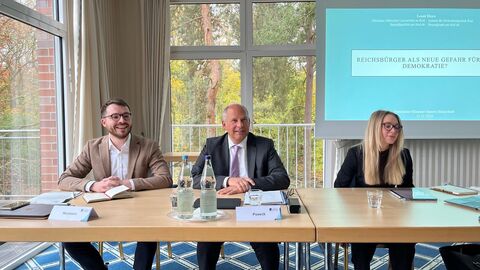 Zwei Männer und eine Frau sitzen an Tisch, im Hintegrund ist eine Präsentation mit dem Titel: Reichsbürger als neue Gefahr für die Demokratie zu sehen.