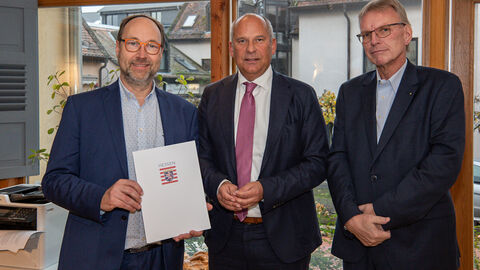Drei Männer stehen nebeneinander. Der linke Mann hält eine Urkunde hoch.