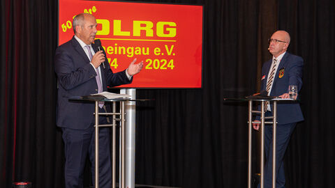 Zwei Männer stehen an Pulten und sprechen miteinander. Im Hintergrund ist das Logo des DLRG