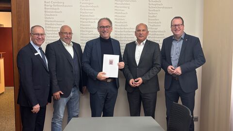 Mehrere Männer stehen im Anzug vor einer Wand mit verschiedenen Stadtnamen