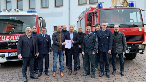 Innenminister Roman Poseck und Bürgermeister Frank Groos halten den Zuwendungsbescheid, umgeben von weiteren Männern, im Hintergrund Feuerwehrwagen und -gebäude.