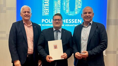 Drei Männer stehen nebeneinander. Der mittlere Mann hält eine Urkunde in der Hand.