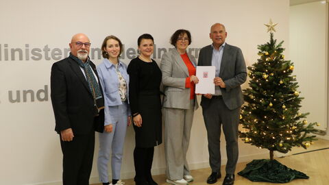 Innenminister Poseck steht mit einer Gruppe Personen neben einem kleinen Weihnachtsbaum. Er überreicht einen Förderbescheid.