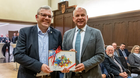 Innenminister Poseck steht neben einem weiteren Mann in einem Saal voller Menschen. Sie halten ein buntes Bild, das als Geschenk verpackt ist, zwischen sich.