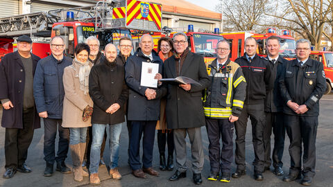 Eine Gruppe Personen steht auf einem Platz bei einer Bescheidübergabe, im Hintergrund sind Feuerwehrfahrzeuge zu erkennen.