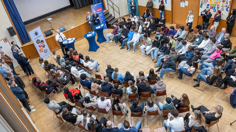 Cops im Dialog Aus der Vogelperspektive aufgenommenes Bild. Im Raum sitzen mehrere Schülerinnen und Schüler, Mehrere Erwachsene stehen.
