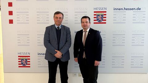 Zwei Männer im Anzug schauen in Kamera. Im Hintergrund steht Hessisches Ministerium des Innern, für Sicherheit und Heimatschutz.