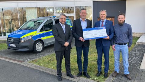Vier Personen stehen vor einem Gebäude, daneben parkt ein Polizeiauto. Die beiden Personen in der Mitte halten gemeinsam einen großen Check.