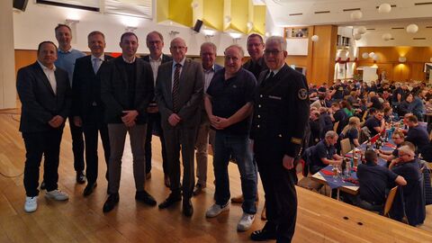 Zehn Männer stehen auf einer Bühne und im Hintergrund sieht man einen Saal gefüllt mit Menschen, die an Tischen sitzen.