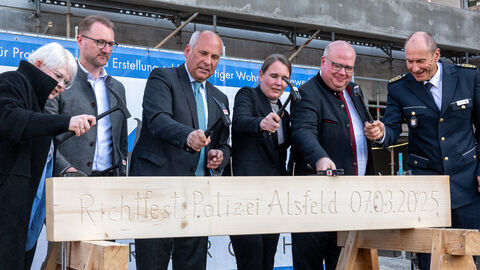 Sechs offiziell gekleidete Personen stehen hinter einem Holzblock und schlagen jeweils mit einem eigenen Hammer Nägel in diesen