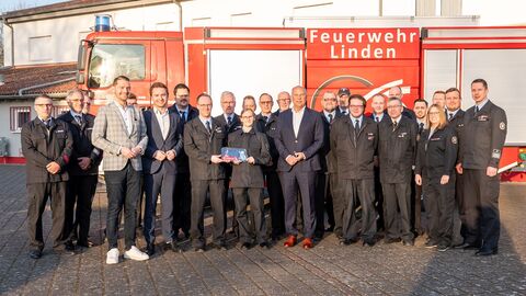Eine größere Personengruppe steht auf einem Platz zum Gruppefoto aufgestellt. Im Hintergrund steht ein Feuerwehrfahrzeug.