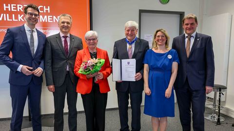 Mehrere Personen stehen zum Gruppenfoto aufgestellt in einem Raum. Eine Dame in der Mitte hält einen Blumenstrauß, der Herr daneben hält eine Urkunde in Händen.