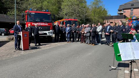 Roman Poseck steht auf einem Parkplatz an einem Rednerpult, hinter ihm stehen zwei Feuerwehrautos und vor ihm eine Gruppe von Menschen, teil in festlichen Uniformen