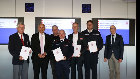 Heimatschutzminister Roman Poseck überreicht Vertretern der Hilfsorganisationen im hessischen Katastrophenschutz in einer Sitzung der Arbeitsgemeinschaft Katastrophenschutz im Krisenzentrum im hessischen Innenministerium Zuwendungsbescheide für die Durchführung der Landesausbildung im Katastrophenschutz Sieben Männer stehen nebeneinander und halten Urkunden in der Hand.