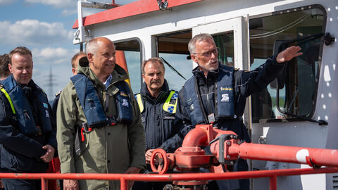 Innenminister Roman Poseck trägt eine Rettungsweste und steht auf einem Schiff, umgeben von vier Polizisten. Einer der Beamten erklärt ihm etwas.
