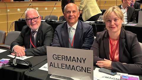 Zwei Männer und eine Frau schauen in Kamera. Auf dem Schreibtisch vor ihnen ist ein Schild mit der Aufschrift "GERMANY ALLEMAGNE"