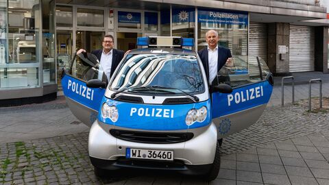 Innenminister Poseck und ein weiterer Mann stehen in der Tür eines kleinen Polizeiwagens vor einem Gebäude.