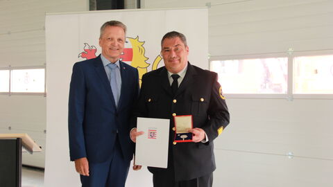 Staatssekretär Martin Rößler und Kreisbrandinspektor Carsten Lauer stehen nebeneinander, Carsten Lauer hält seine Urkunde und das Ansteckkreuz in Händen.