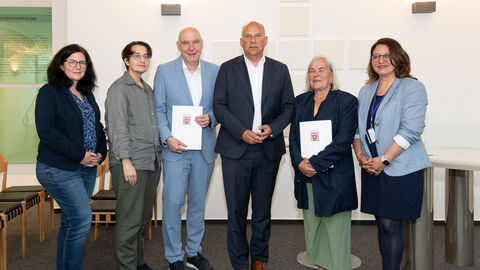Sechs Männer und Frauen stehen nebeneinander und halten ein Dokument in der Hand.