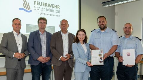 Sechs Frauen und Männer stehen vor einer Projektorwand und halten ein Dokument hoch.