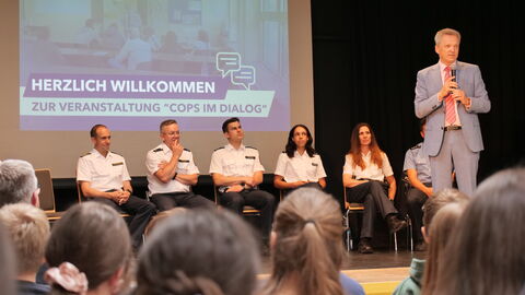 Cops im Dialog Innenstaatssekretär Rößler steht auf einer Bühne und spricht. Im Hintergrund sitzen Polizistinnen und Polizisten.