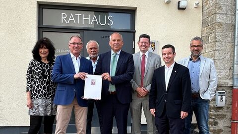 Eine Frau und mehrere Männer stehen vor einem Gebäude mit Aufschrift "Rathaus" und lächeln in Kamera. Zwei der Männer halten einen Zuwendungsbescheid in den Händen.