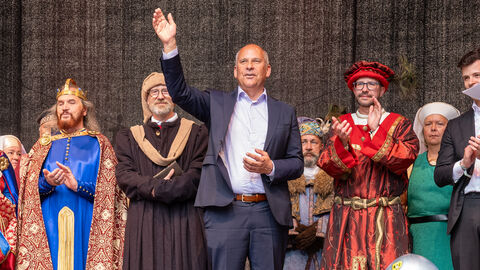 Roman Poseck steht auf der Bühne und winkt ins Publikum. Um ihn herum zahlreiche Personen in historischen Gewändern. 