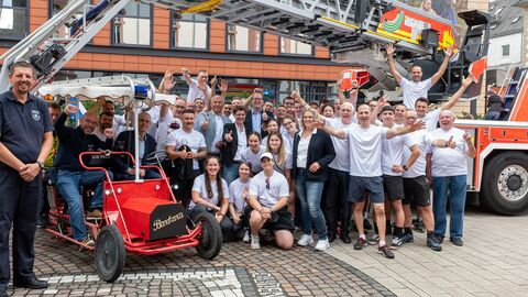 Viele Menschen in weißen T-Shirts lachen in die Kamera. Im Hintergrund sieht man ein Feuerwehrauto.
