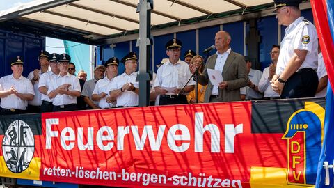 Mehrere Männer, darunter Feuerwehrleute, stehen auf einer Bühne. Ein Mann hält eine Rede. Auf einem Banner steht "Feuerwehr- retten-löschen-bergen-schützen"